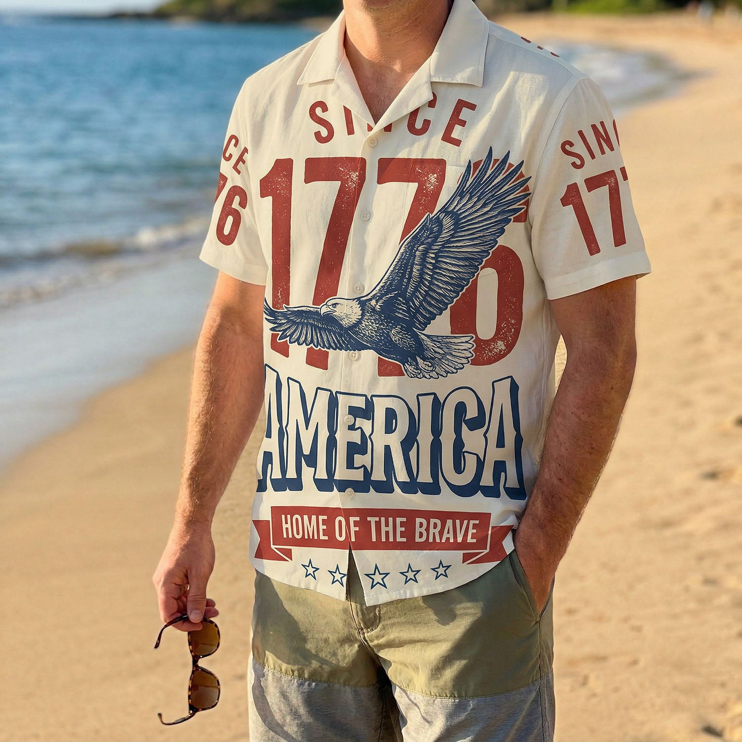 A man wearing the cream 1776 Home of the Brave shirt on a sunny beach, showcasing a relaxed patriotic summer look.